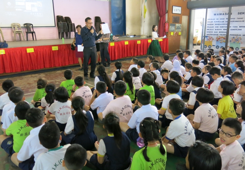 新亚小学校长马绅涵向四年级学生讲解马来文课本介绍爪夷文单元的问卷内容，以让学生把问卷带回给家长填选赞成或不赞成。