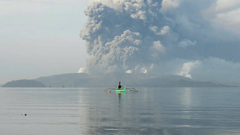 A youth living at the foot of Taal volcano rides an outrigger canoe while the volcano spews ash. Photo courtesy: AFP