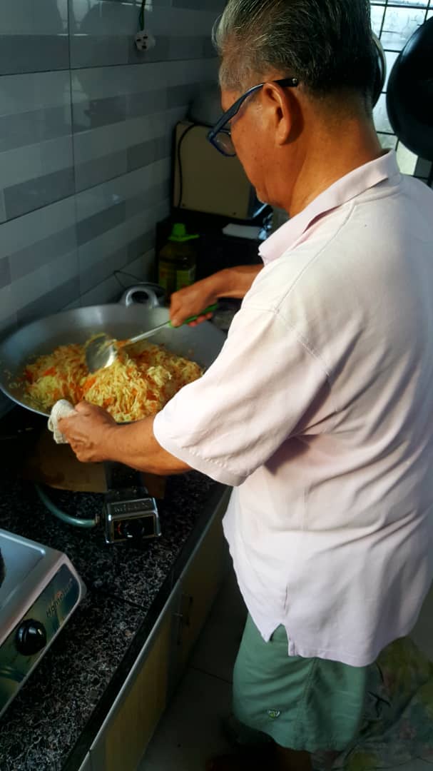 梁秀玲的父亲在家协助炒菜，减轻女儿的工作量。