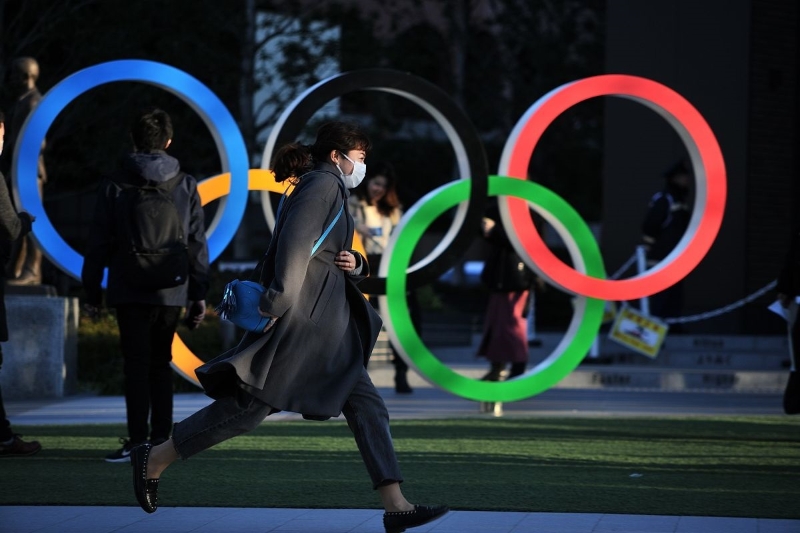 东京奥运会能不能如期主办，早已成为了国际话题。图示一名戴着口罩的女子，行色匆匆地走过东京市一处奥运五环标志碑的画面。