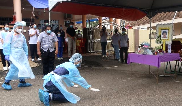 Clad in a quarantine outfit, Tung Ming Shan kneels down to pay his last respect to his late mother. Also clad in quarantine outfit, the civil defense officer is standing beside him. SIN CHEW DAILY Clad in a quarantine outfit, Tung Ming Shan kneels down to pay his last respect to his late mother. Also clad in quarantine outfit, the civil defense officer is standing beside him. SIN CHEW DAILY