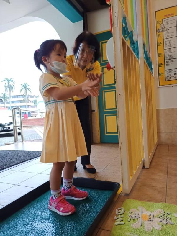 女生站在塑胶盆内替鞋子消毒，一旁的雷丽茵教导她如何利用搓手液洗手。