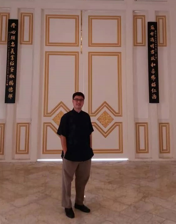 杨金荣在新山大王宫华族礼堂前留影，背后的木制牌匾正是他题字及雕刻的作品。