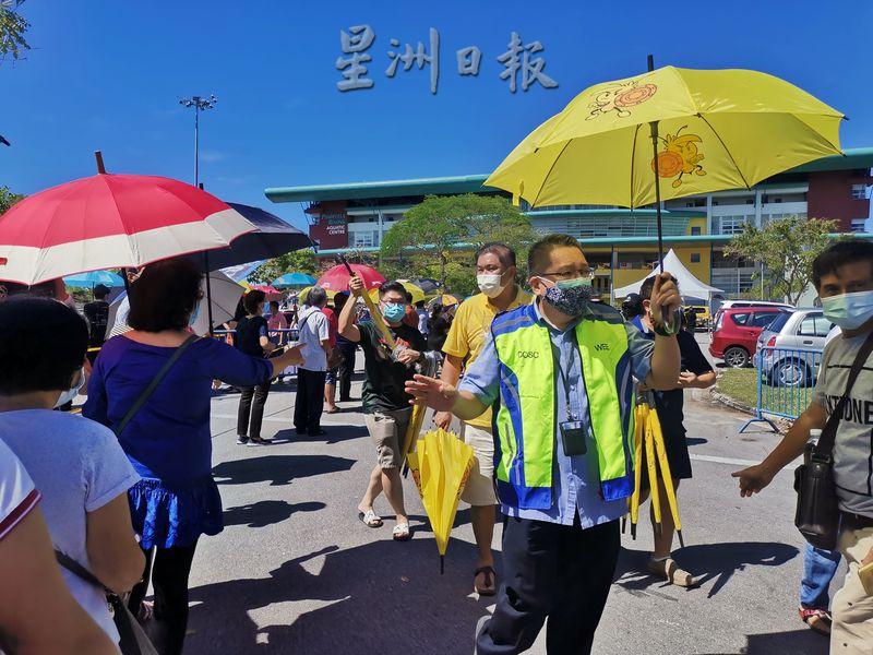 古晋南市市长拿督黄鸿圣在接种中心外派发雨伞给在烈日下排队的民众。
